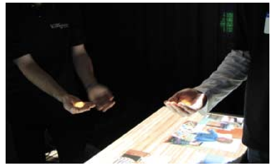 Image of two people in front of a table, a projector rendering an image on the palm of their hands